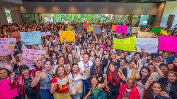 Multitud de mujeres de distintas edades y vestimentas reunidas en una asamblea al aire libre en Morelia, con pancartas y banderas, escuchando a Carlos Torres Piña.