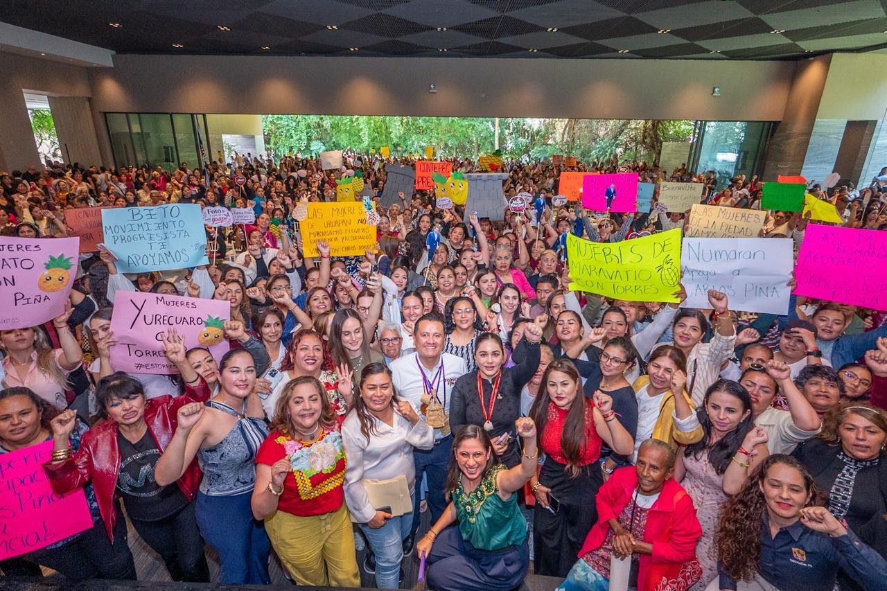 Multitud de mujeres de distintas edades y vestimentas reunidas en una asamblea al aire libre en Morelia, con pancartas y banderas, escuchando a Carlos Torres Piña.