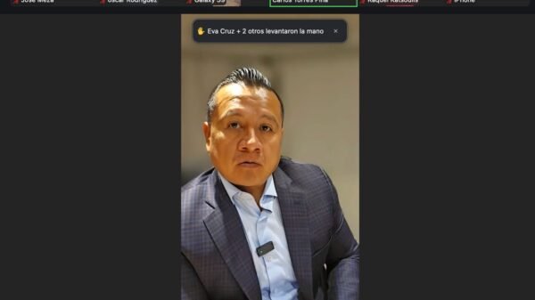 Carlos Torres Piña hablando frente a un mural con símbolos de Michoacán y una bandera mexicana, rodeado de migrantes y representantes comunitarios