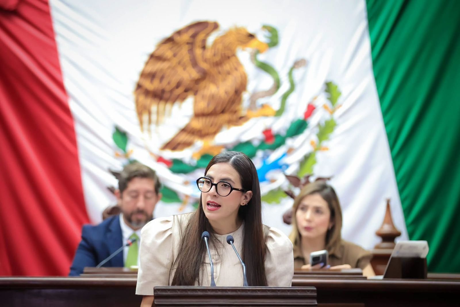 Diputada Giulianna Bugarini hablando ante micrófonos en el Congreso de Michoacán, con fondo del escudo estatal