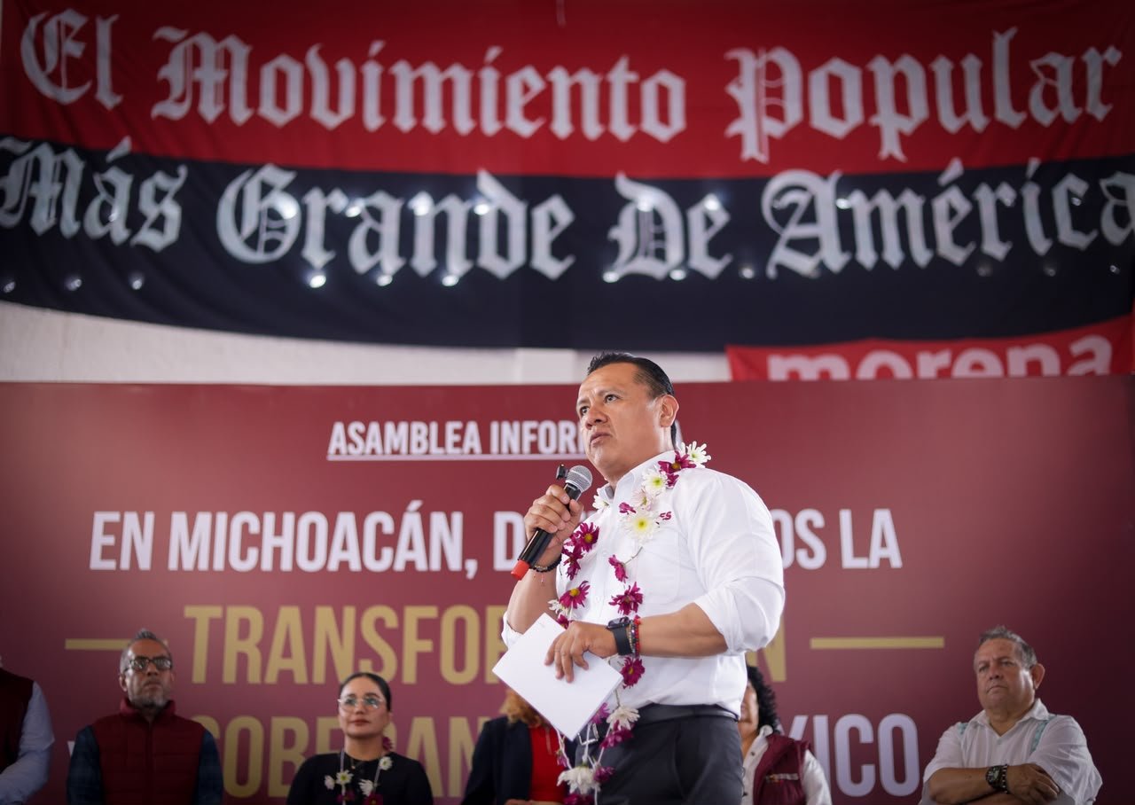 Carlos Torres Piña dialogando con ciudadanos de distintos sectores en un municipio rural de Michoacán, bajo un cielo despejado y con letreros comunitarios visibles