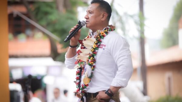 Carlos Torres Piña hablando ante micrófono en evento público en Morelia, Michoacán, con bandera mexicana al fondo