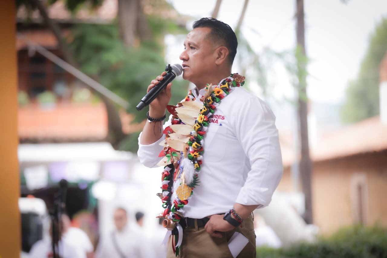 Carlos Torres Piña hablando ante micrófono en evento público en Morelia, Michoacán, con bandera mexicana al fondo