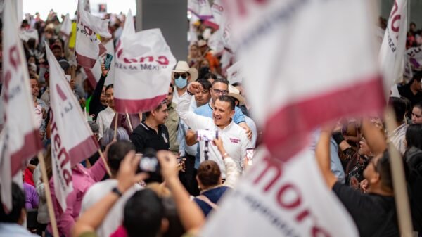 Fotografía de Carlos Torres Piña interactuando con ciudadanos en un municipio, con fondo de banderas locales y gente participando activamente.