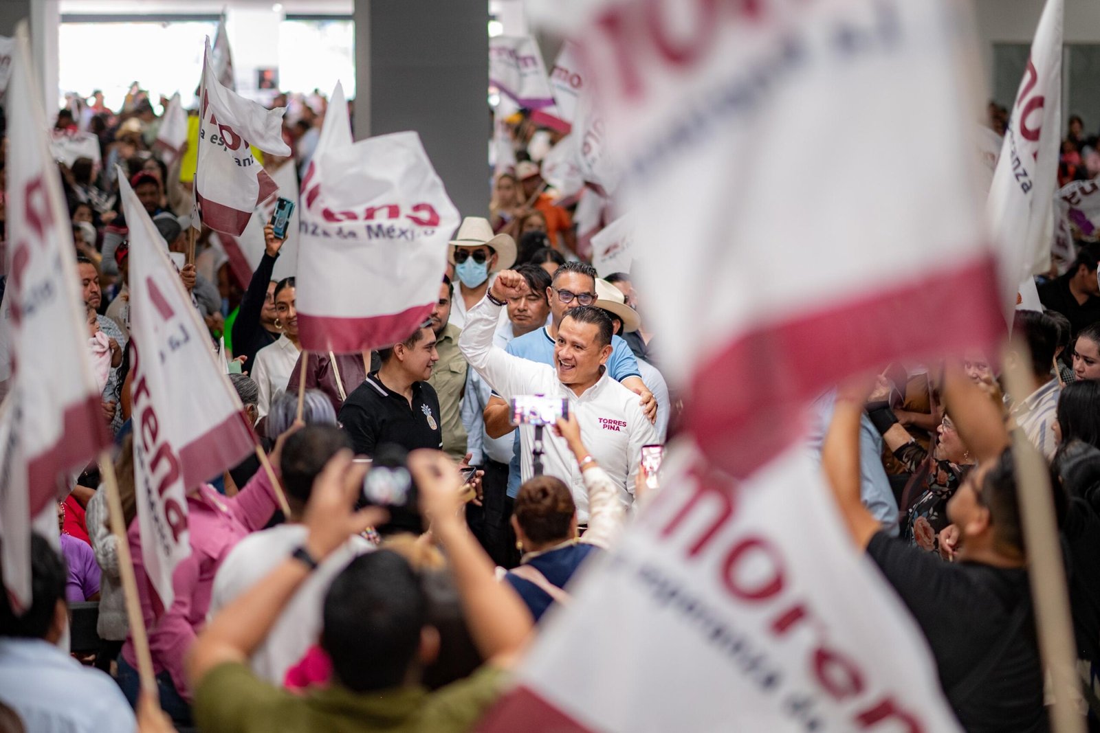 Fotografía de Carlos Torres Piña interactuando con ciudadanos en un municipio, con fondo de banderas locales y gente participando activamente.