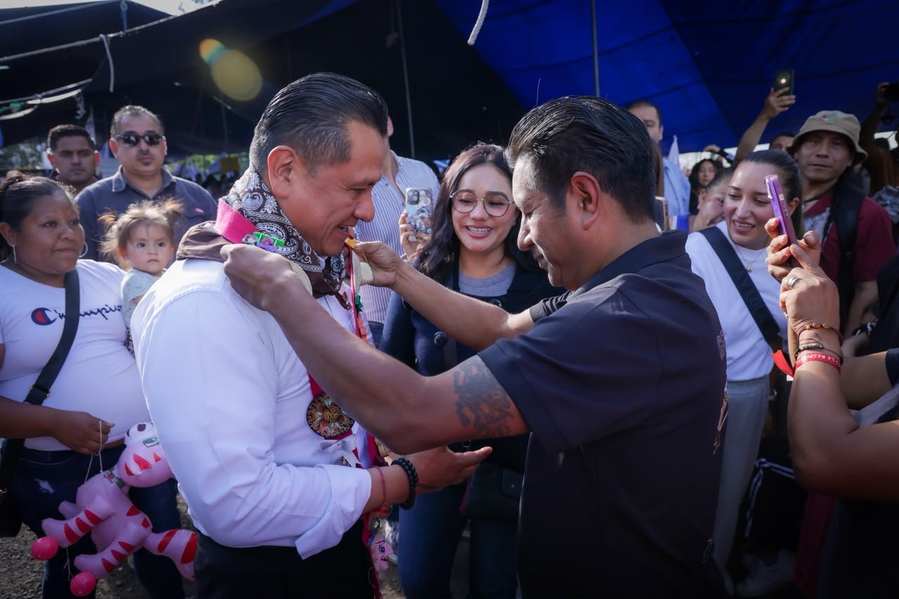 Fotografía de Carlos Torres Piña conversando con ciudadanos en un espacio público, con micrófono y libreta, durante uno de sus recorridos por el estado