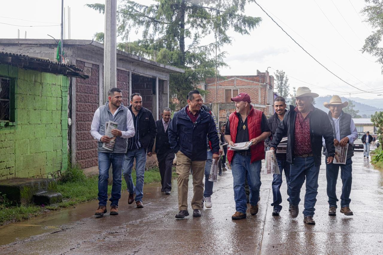 Fotografía de Carlos Torres Piña conversando con habitantes de un municipio michoacano, en entorno rural, con mapa de Michoacán visible de fondo