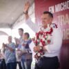 Carlos Torres Piña hablando ante vecinos de Tiripetío en la plaza principal, con bandera de Morena visible y público diverso de todas las edades
