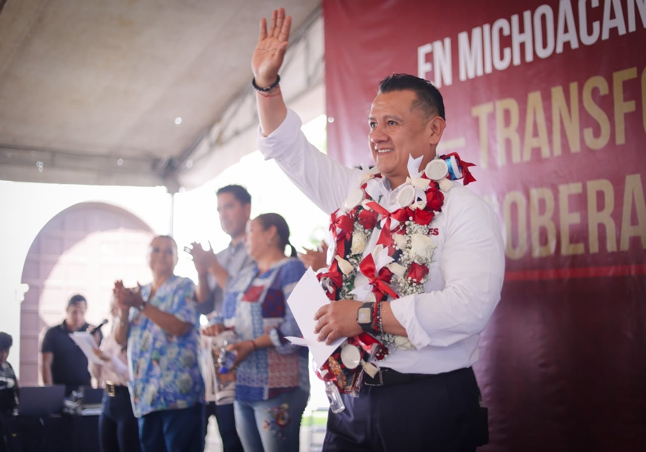 Carlos Torres Piña hablando ante vecinos de Tiripetío en la plaza principal, con bandera de Morena visible y público diverso de todas las edades