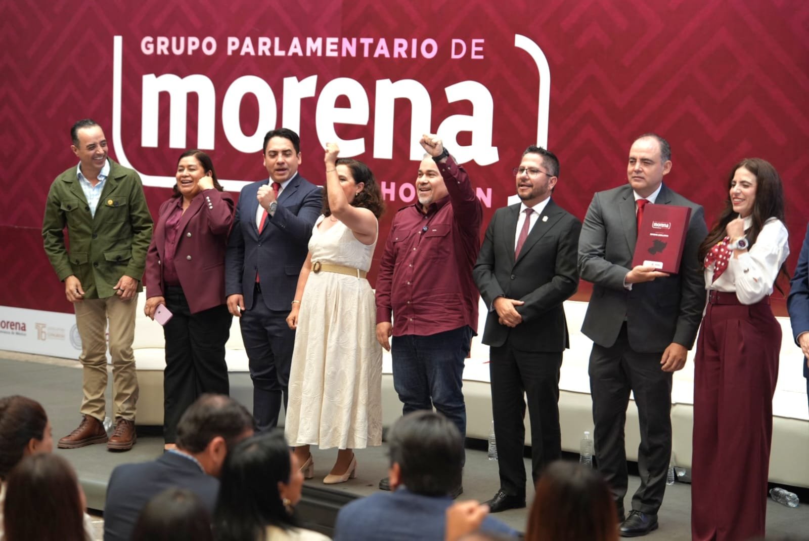 Retrato formal de la diputada Belinda Iturbide Díaz frente a un fondo con los colores de la bandera de Michoacán y símbolos de salud, educación y justicia
